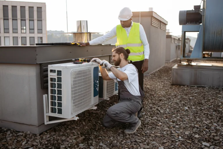 Caucasian male builder in overalls crouching and repairing air conditioner while using screwdriver. Black supervisor wearing white helmet standing and watching work of partner on roof of factory.