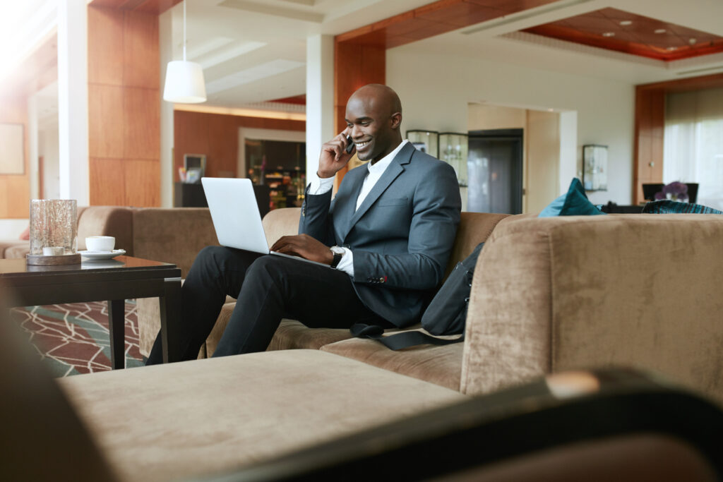 A man in a suit talks on the phone with a laptop in his lap. He is in a hotel lobby. He is smiling.
