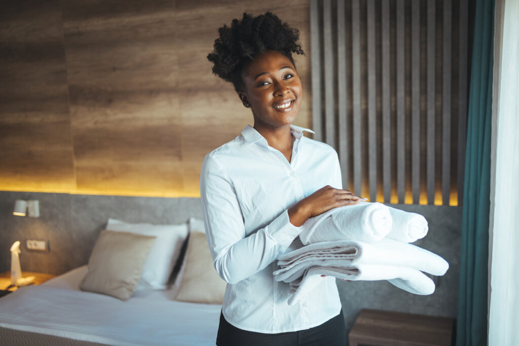 Woman with curly hair and dressed in maid uniform of a luxurious hotel arranges the towels of future guests and enjoys the moment smiling for the camera. Maid making bed in hotel room.