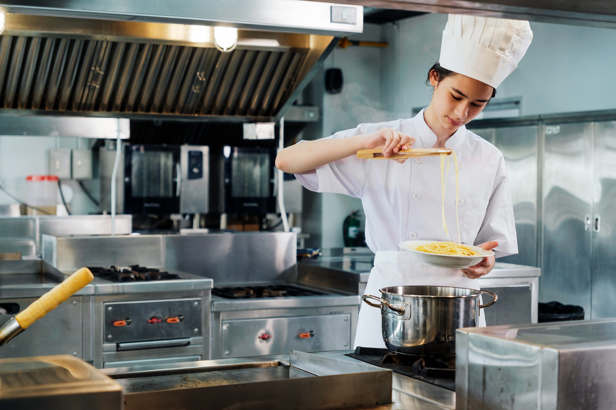 Professional chef in uniform plates pasta in stainless industrial kitchen, sprinkling seasoning. Scene shows skills, technique, training, hospitality, heat, stove work, fast-paced restaurant service. kitchen equipment