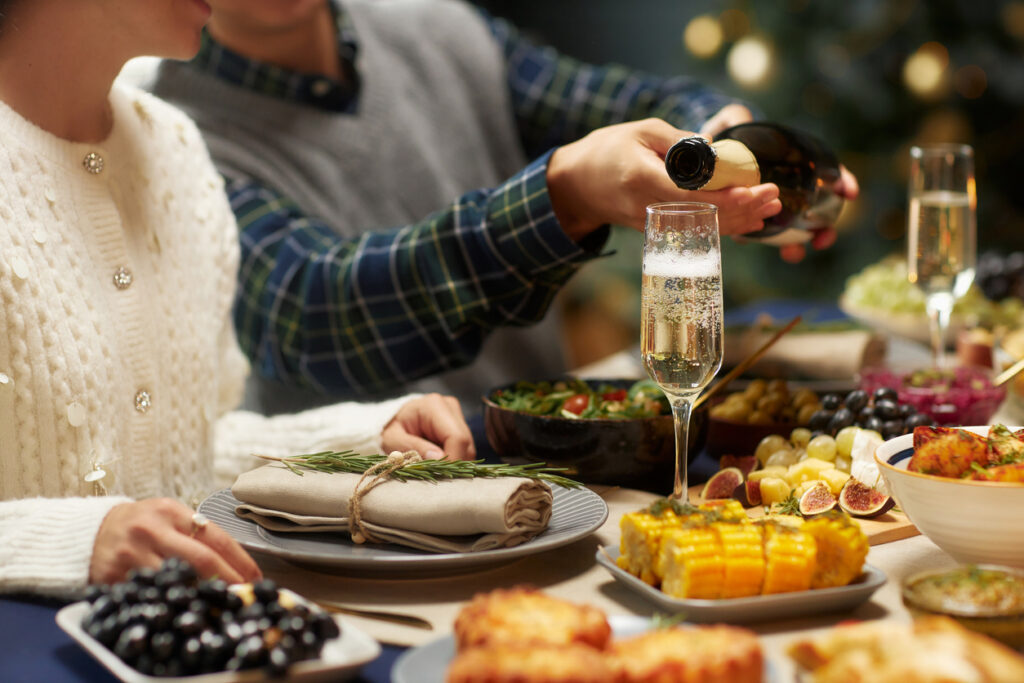 Cropped shot of man pouring sparkling champagne into glass helping female friend while sitting at dining table during festive dinner while celebrating Christmas, copy space