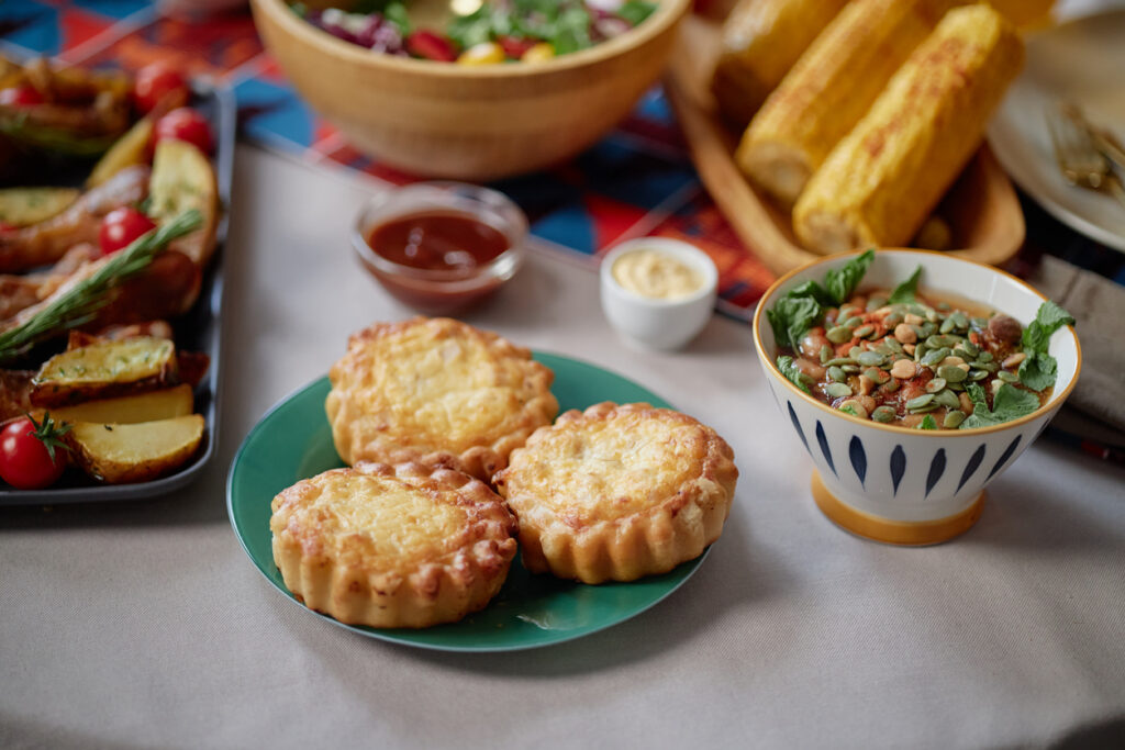 Closeup showing traditional Kwanzaa meal with baked cheese tarts on plate, fresh salad with seeds in bowl, roasted vegetables and corn on table, no people visible